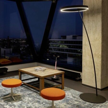 Designer-Stehleuchte mit gebogenem Stab und Ringbeleuchtung in einer Loft-Wohnung bei Nacht; im Vordergrund ein schlichter Couchtisch aus Holz und 2 Hocker mit orangefarbenem Bezug